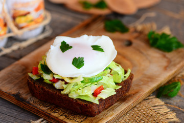 Simple vegetable salad and poached egg sandwich. Poached egg on rye bread slice with fresh vegetable salad on a wooden board. Poached eggs for breakfast weight loss. Rustic style. Closeup