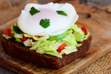 Home egg poached sandwich. Egg poached on rye bread slice with fresh cabbage, cucumber, pepper and parsley. Healthy food. Wooden background. Rustic style. Closeup