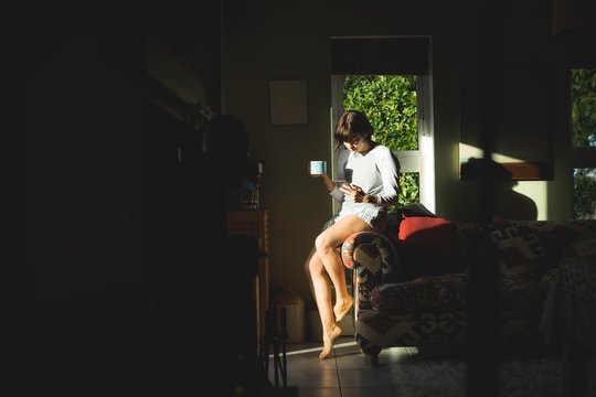 Woman having coffee while using mobile phone in living room