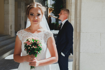 Wedding blonde couple is walking in the old city. Groom is standing near the ancient stone walls columns. Bride in lace satin dress is holding pink rose bouquet. Sunny love story in the medieval town.