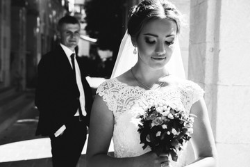 Wedding blonde couple is walking in the old city. Groom is looking at the bride in lace satin dress with pink rose bouquet. Sunny love story near the ancient stone walls columns. Black and white photo