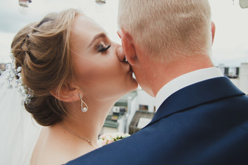 Wedding blonde couple is embracing in the ancient cafe on the roof. Groom and bride in lace satin dress are kissing. Close up portrait of a bride with professional makeup and long lashes.