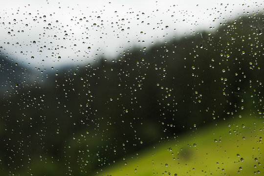 Raindrops of rain on glass of window of cottage at countryside area. View at blurry green forest and mountain hills behind glass. Horizontal color photography.