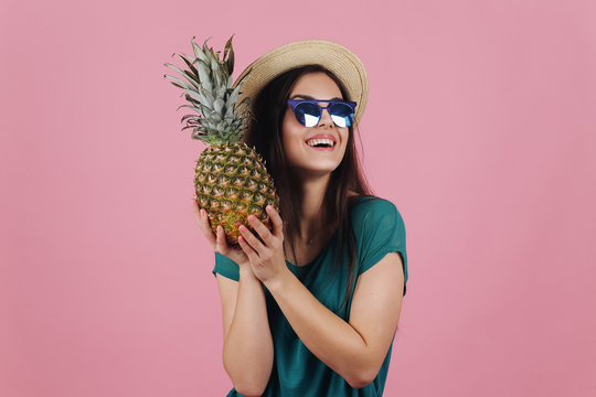 Laughing girl in sunglasses and a hat stands with a pineapple in her arms