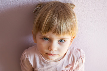 Sad little girl with blond hair is leaning against the wall in a sunny room at home. She looks at the viewer. Top view, close up.