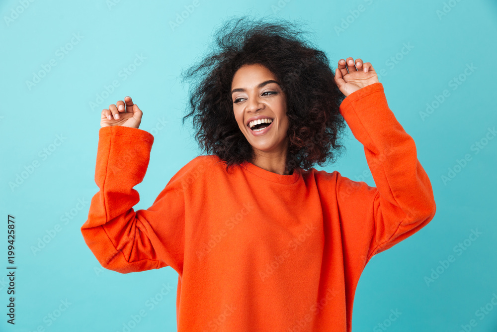 Wall mural Happy portrait of excited woman in red shirt looking on camera with smile and raising hands, isolated over blue background - Wall murals