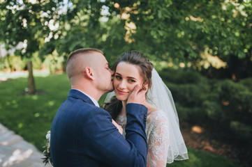 A young groom kisses a beautiful bride in a lace dress in a green park. Portrait of a newlywed couple.