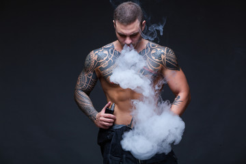 Vaper. The man with a muscular naked torso with tattoos smoke an electronic cigarette on the dark background