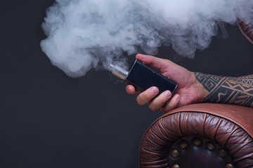 Hand close up. Vaper. The man with tattoos sits on a leather sofa smoke an electronic cigarette on the dark background