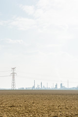 View of a plowed land with a transmission tower, high-voltage power lines and the silhouettes of the smoking chimneys of a refinery on the horizon under a pale sunlight.