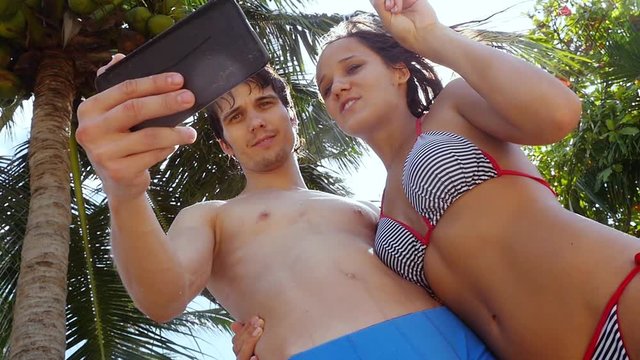 Romantic cheerful couple in bikini taking selfie on cell smart phone outdoors under palm trees on the sun and sun flare on the background. slow motion. 1920x1080