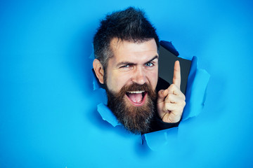 Bearded man making hole in paper. View of male face through hole in blue paper. Bearded man looking through hole and show the finger. Bearded man making hole in paper and points up.