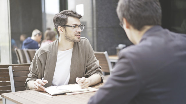 Two friends are having a conversation at table of the cafe outdoors. A man dressed in a jacket wearing eyeglasses is listening carefully to his friend talking