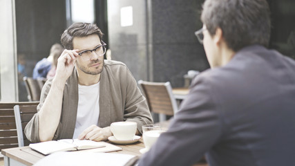 Two friends are having a conversation at table of the cafe outdoors. A man dressed in a jacket...