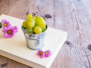 Grüne Weintrauben und rosa Blüten auf einem Buch auf einem grauen Holztisch.  Textfreiraum.