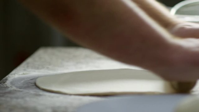Close up hands of unrecognizable restaurant chef rolling out dough on floured surface and forming circle for future pizza