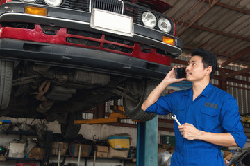 Asian mechanic speaking on phone under the car to repair the engine with work board in hand, japanese mechanic portrait style, mechanic maintenance working under car