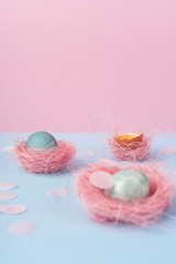Yolk of broken egg in eggshell and two blue eggs decorated with pink sisal nest and pink confetti on the minimalist  pink and blue background