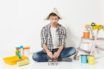 Fun crazy young man in newspaper hat with supermarket grocery push carts for shopping. Instruments for renovation apartment isolated on white background. Wallpaper painting tools. Repair home concept.