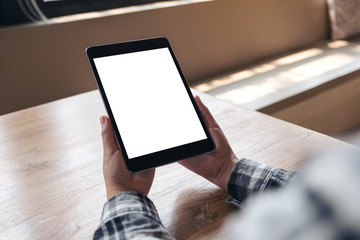 Mockup image of hands holding black tablet pc with blank white desktop screen on wooden table