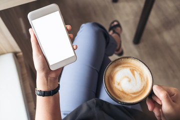 Top view mockup image of a woman's hand holding white mobile phone with blank desktop screen while drinking coffee in cafe