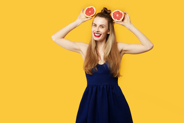 Young woman on a yellow background and a blue dress is holding a cut orange in her hands. Concept of healthy nutrition and sports.  Colour obsession concept.  Minimalistic style. Stylish Trendy
