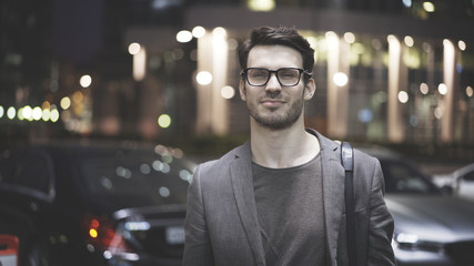 A closeup of a bearded man in jacket wearing glasses on the street at night. A hipster in a night time in a big city looking at the camera with a delight smile