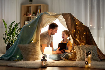family, hygge and technology concept - happy father and little daughter with tablet pc computer in kids tent at night at home