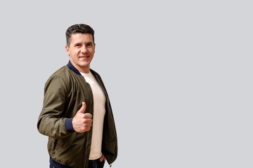 Attractive smiling middle-aged man in a green  jacket stands on a gray background and makes a gesture of thumbs up.