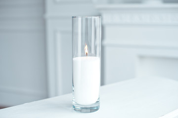 A burning candle in tall glass vase flask standing on the table indoor.
