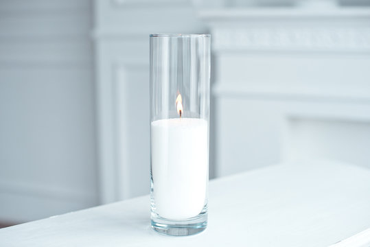 A burning candle in tall glass vase flask standing on the table indoor.
