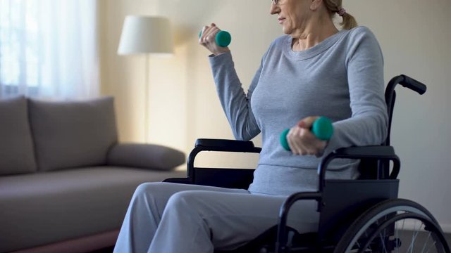 Athletic grandmother in wheelchair lifting dumbbells at home, keeping fit, sport