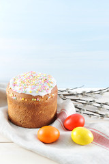 Easter cake on cloth napkin. Around the chicken eggs are red, yellow and orange. In the frame of the willow branch. Light wooden background. Close-up. Vertical orientation of the frame.