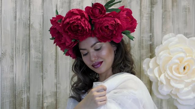 Girl posing in front of camera. young woman in a wreath of scarlet peonies on her head, dark long curly hair descends on the shallow shoulders.