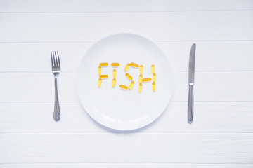 Top view white plate with Fish word lettering by cod Liver Oil Capsules, Omega 3 served with knife and fork on the white wooden table. Healthy living concept. Selective focus, space for text.