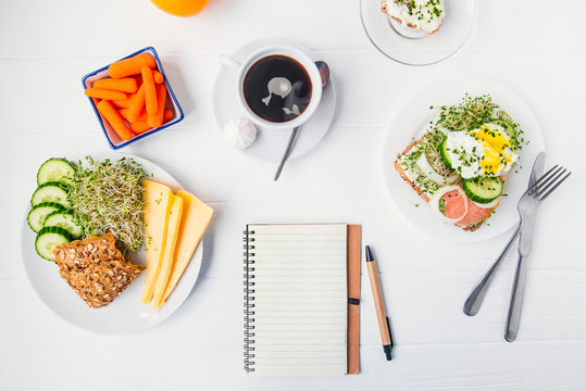 Top view planning notebook with copy space and healthy breakfast. Sandwich with egg benedict and smoked salmon, plate with cheese, sprout micro greens and cucumber, carrots and coffee on wooden table.