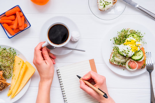 Morning habits of successful people. Day planning and healthy eating. Woman drink coffee and writing in notebook on the served for breakfast white wooden table. Top view, Selective focus.