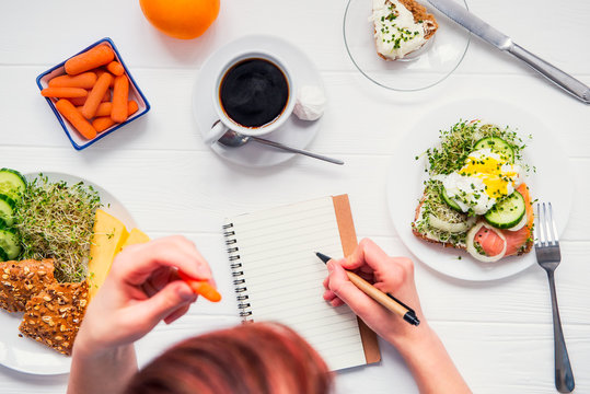 Morning habits of successful people. Day planning and healthy meal. Woman eating carrot and writing in notebook on the served for breakfast white wooden table. Top view, Selective focus.