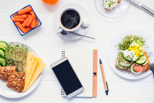 Mobile phone, planning notebook and healthy breakfast. Day diet planning and healthy eating concept. Sandwich with egg benedict, other snacks and coffee on wooden table. Selective focus, copy space.
