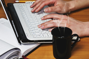 A cup of hot coffee on the table near the laptop on which is printed female fingers closeup