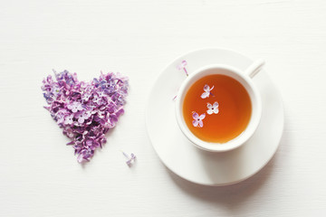 Cup of tea on a saucer and heart from flowers of a lilacin on a white board, top view
