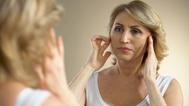 Unhappy aged woman looking in mirror at home, touching her face, aging process