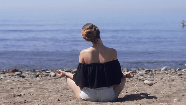 Young woman makes meditation in lotus pose on sea / ocean beach, harmony and contemplation. Beautiful girl practicing yoga at sea resort at her vacation. Life style. Young woman relaxing outdoors.