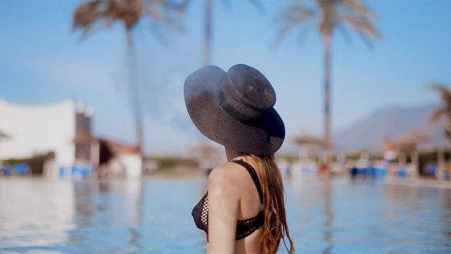 Blonde girl smoking electronic cigarette near sweaming pool in a hat and sun glasses. Beackgroung palm, sun, pool.Young beautiful woman smoking ( vaping ) e-cigarette on a beach. Vacation. Tourism.