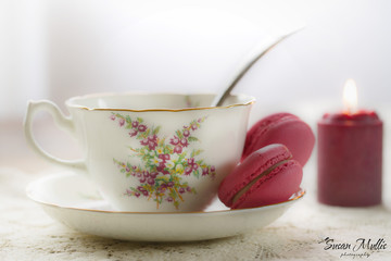 Porcelain cup and saucer with red raspberry macaroon cookies with burning candle in background