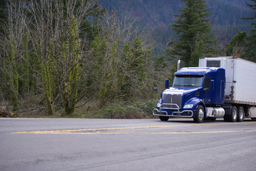Fototapeta na wymiar Dark blue big rig semi truck with refrigeration semi truck transporting goods in wide road with forest trees background
