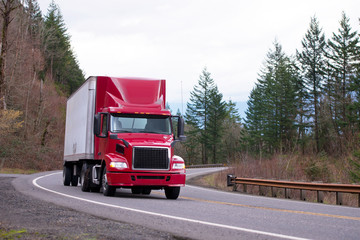 Red big rig day cab semi truck with semi trailer running on winding road with trees and safety fence