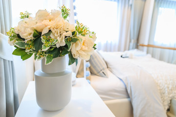 A bouquet of artificial flower in a pot with bed and blinds background.