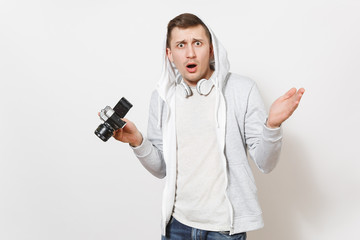 Young handsome surprised male student in t-shirt and light sweatshirt with hood with headphones holds retro camera and spreads his hands isolated on white background. Concept of photography, hobby
