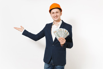 Smiling businessman in dark suit, protective orange hardhat holding bundle of dollars, cash money and pointing hand aside on copy space isolated on white background. Male worker for advertisement.